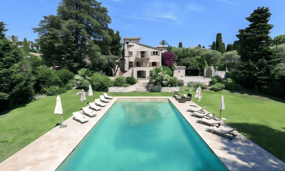 Aerial view of a turquoise rectangular pool at Villa Cap d’Antibes, lined with sun loungers and white umbrellas, framed by a manicured lawn and a stone villa facade