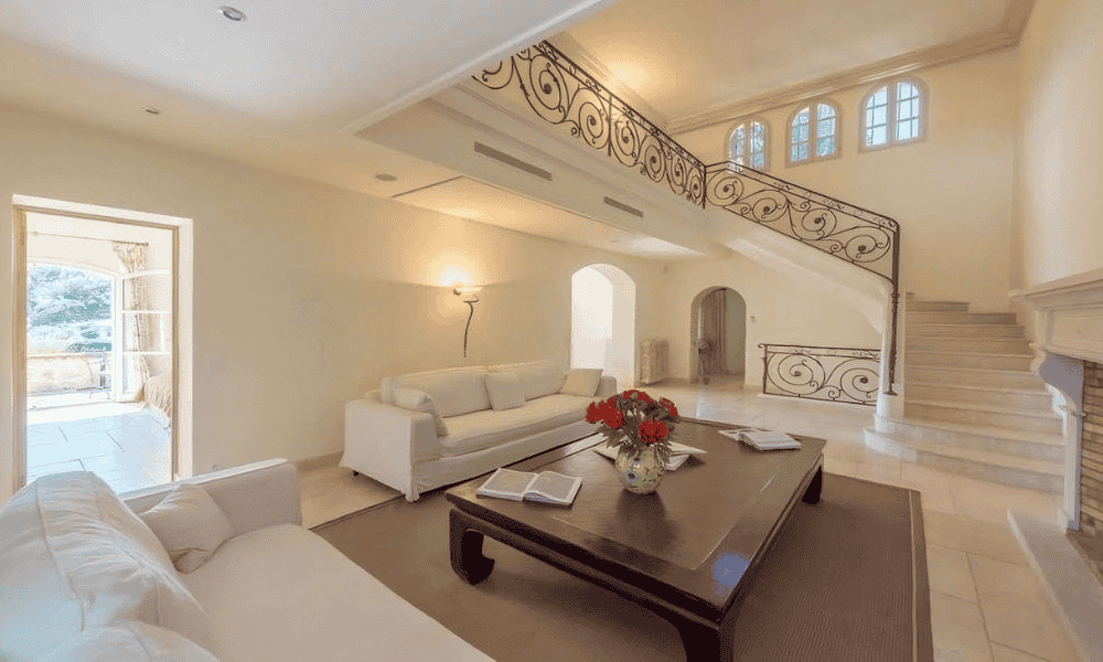 Double-height living room at Villa Cap d’Antibes with white sofas, dark wood coffee table topped with red flowers, ornate wrought-iron staircase, and fireplace