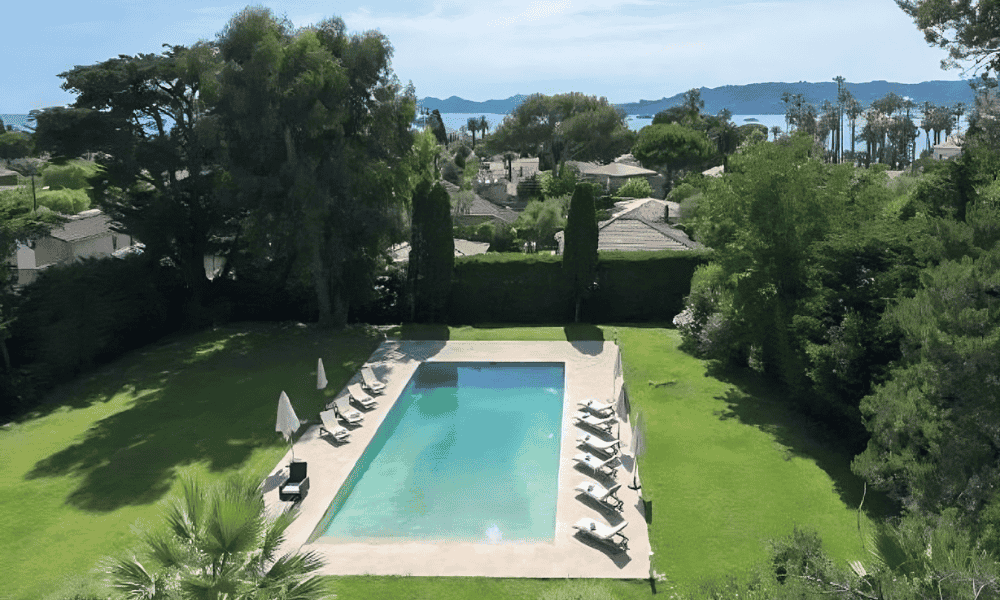 High-angle view of the rectangular pool and surrounding lawn at Villa Cap d’Antibes, with sun loungers and umbrellas, overlooking tiled rooftops and the Mediterranean Sea beyond