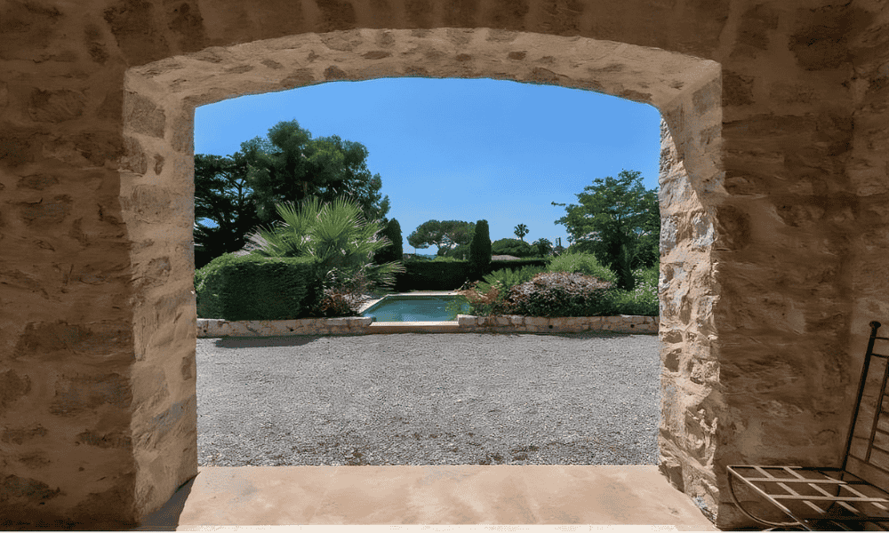 View through a rustic stone archway framing the pool and hedged garden at Villa Cap d’Antibes under a clear blue sky