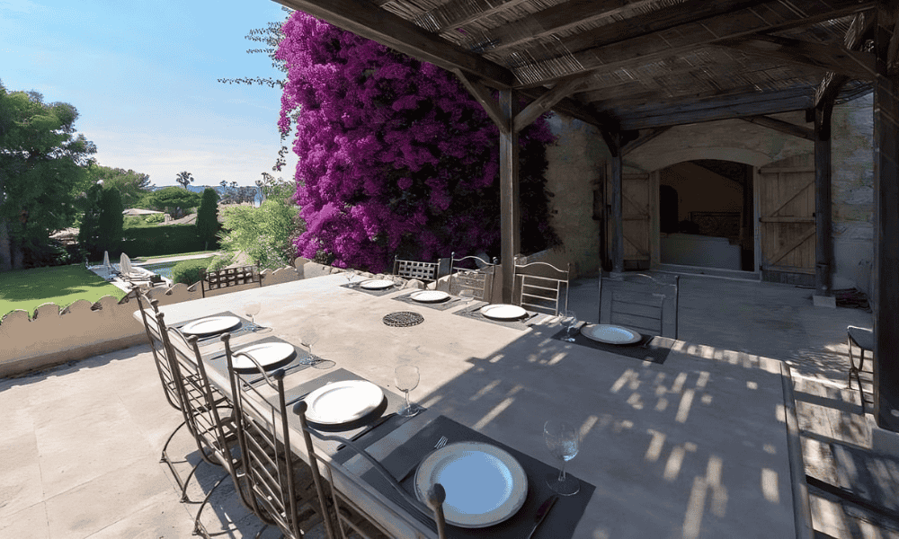 Shaded outdoor dining terrace at Villa Cap d’Antibes under a wooden pergola, set with plates and glassware beside vibrant purple bougainvillea