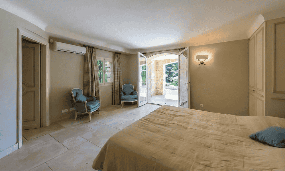 Bright bedroom suite at Villa Cap d’Antibes featuring a queen bed with beige linens, two blue upholstered armchairs, and open French doors leading to a sunlit courtyard