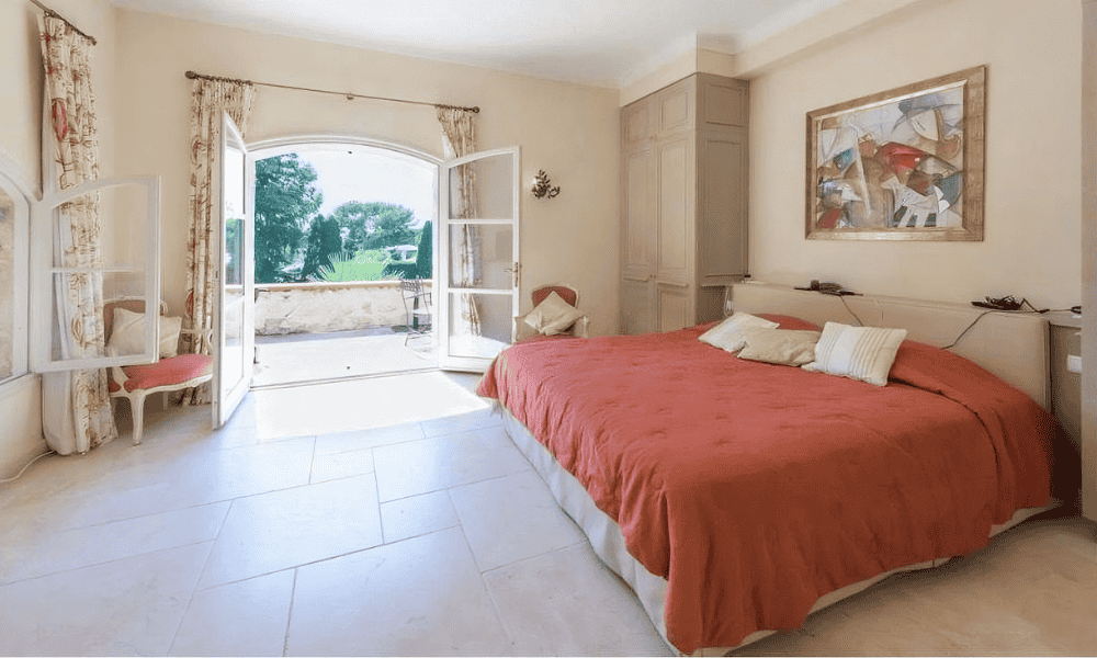 Bedroom at Villa Cap d’Antibes with red duvet on king-size bed, abstract artwork above, and open French doors leading to a sunny terrace