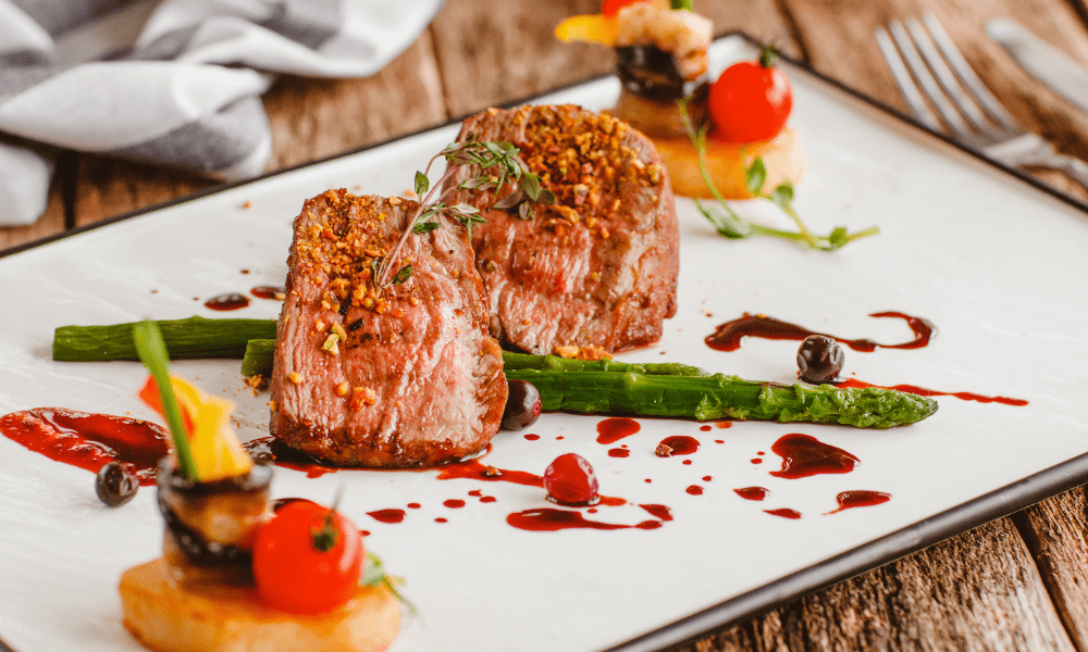 Seared beef medallions garnished with crushed nuts and thyme, accompanied by grilled asparagus, cherry tomatoes, berries, and red wine reduction on a white plate