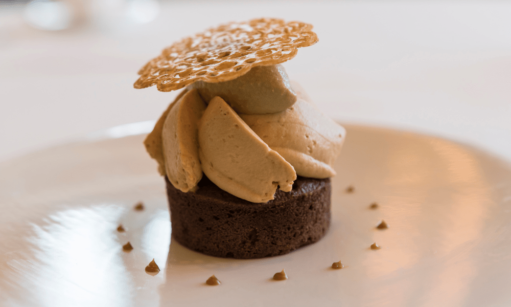 Mini chocolate cake topped with hazelnut cream swirls and a delicate caramel lace tuile, served with dots of caramel sauce on a white plate