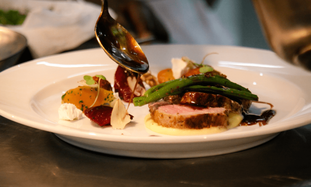 Chef spooning rich brown sauce over sliced roast meat on creamy mashed potatoes, garnished with asparagus spears and seasonal vegetables