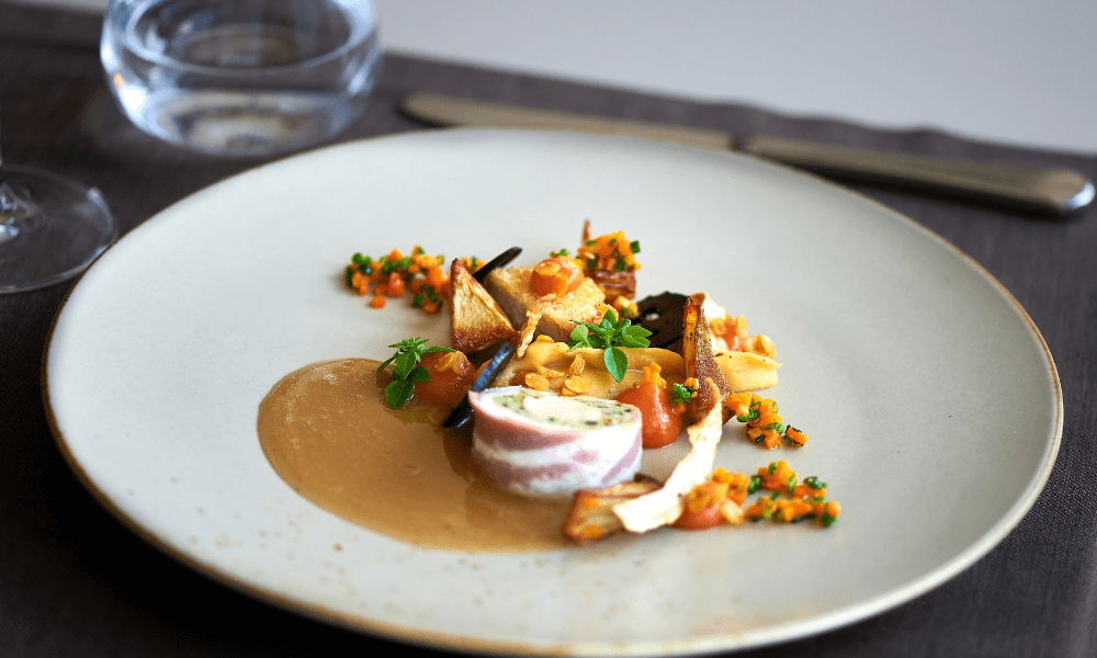 Villa Hotels gourmet bacon-wrapped roulade with creamy herb filling, served on a bed of rich brown sauce, accompanied by artichoke crisps, carrot succotash, and fresh microgreens