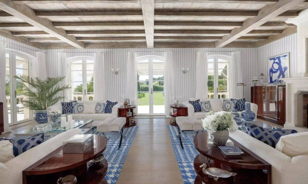 Bright villa living room with exposed wooden beam ceiling, white sofas accented by blue patterned cushions, glass coffee table, and garden view through French doors