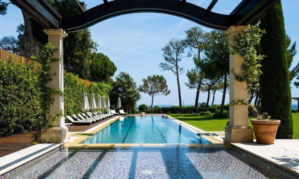 Outdoor infinity pool with sun loungers under a pergola, set amid lush greenery overlooking the sea