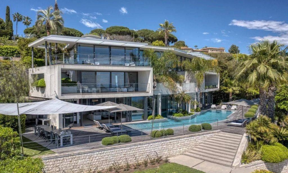 Aerial exterior view of a contemporary luxury villa with floor-to-ceiling glass, infinity pool, and Mediterranean hillside backdrop
