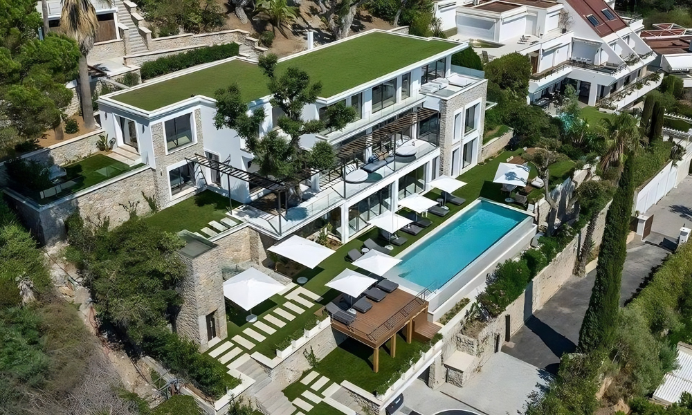 Aerial view of a hillside villa with green rooftop terrace, infinity pool, and white umbrellas overlooking coastal landscape